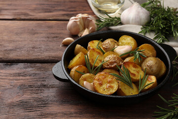 Delicious baked potatoes with rosemary in frying pan on wooden table. Space for text