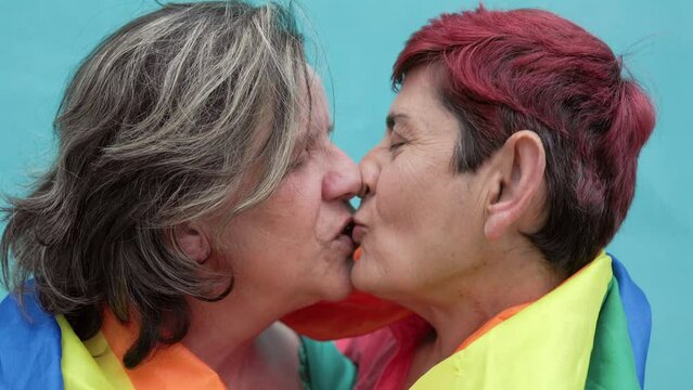 Happy gay senior lesbian couple kissing wearing lgbt rainbow flag outdoors - Diversity family love