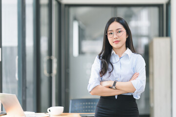 A portrait of a smiling, young, beautiful, professional, and confident millennial Asian businesswoman standing at a modern office.