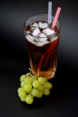 A tall glass of fruit juice with straws and ice on a black background, next to a bunch of ripe green grapes.