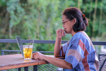 Fototapeta premium Asian woman sitting at her home balcony working on her laptop computer with a glass of lemon tea on the table