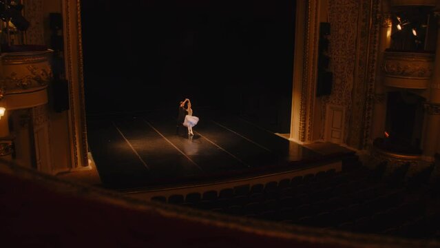 Establishing shot of ballet dancers preparing theatrical dance performance. Man and woman practice choreography on classic theater stage. Classical ballet dance. Rows of seats. Dramatic lighting.