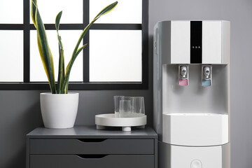 Modern water cooler, drawer, glasses and houseplant near window