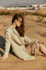 a woman in a stylish jacket and dark glasses sits on the sand relaxing