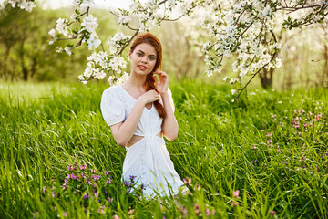 a young woman in a light summer dress stands near a tree with flowers and smiles looking at the camera