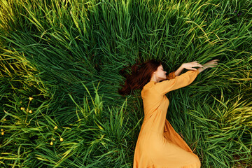 an elegant woman lies in the tall green grass in a long summer orange dress and relaxed posing with...