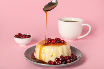Pouring of caramel syrup from spoon onto delicious pudding and cranberries on pink background