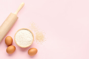 Bowl with wheat flour, sesame seeds, rolling pin and eggs on pink background