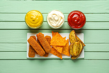 Composition with bowls of ketchup, mayonnaise, mustard and snacks on green wooden table