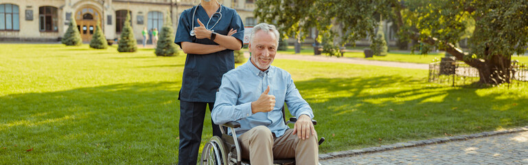 Joyful senior man, recovering patient in a wheelchair smiling at camera, showing thumbs up together with a nurse wearing face shield and mask on a summer day outdoors