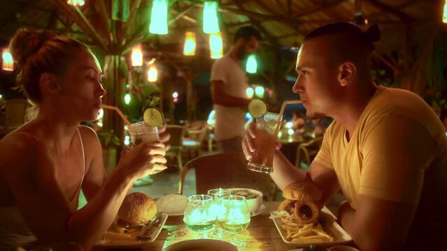 The first date couple in an open outdoor cafe, enjoying talking, laughter, fries, burgers, and cocktails. Candles create cozy atmosphere as colorful cafe lights blur in the background. Summer evening