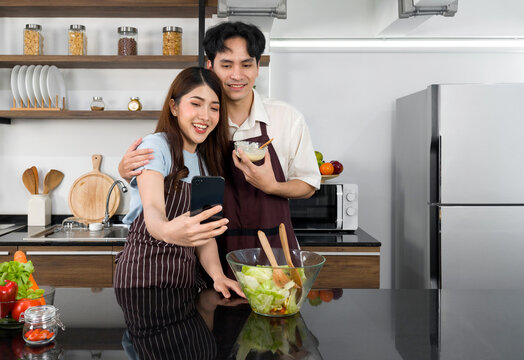 Asian couple spend time together in the kitchen. Young woman take selfie photo with mobile phone while her boyfriend holding salad dressing bowl. Modern lifestyle people relationship and activity