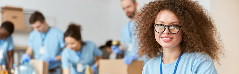 Attractive young woman, volunteer in blue uniform using tablet pc and smiling at camera while sitting indoors. Team sorting, packing items in the background