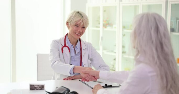 Smiling female doctor shaking hands with mature woman at clinic meeting. Elderly patient making health insurance deal
