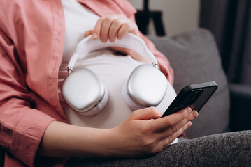Fototapeta premium CLose up of young pregnant woman applying headphones on belly headphones enjoying relaxing music, listening to playlist from smartphone, radio. Peaceful expectant mom taking tranquility sound therapy