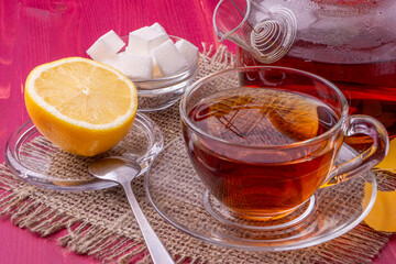 A teapot and a glass cup with brewing black tea. Next to the sliced lemon and lump sugar. Beautiful bright wooden background.