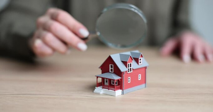 Closeup of business woman hand holding magnifying glass over house model over table. Expert assessment of house by realtor
