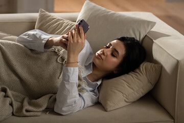 A newly woken woman checks her missed notifications on her smartphone. A young woman is lying on the couch and browsing social media on her smartphone.