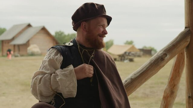 Bearded village squire laughs and chats holding bottle of magic potion in bosom. Positive medieval man rests on sunny day closeup on blurred background
