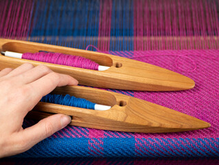 Hand takes a wooden boat shuttle with blue yarn to make a weaving project. Hand loom work