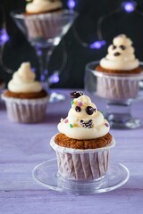Carrot cupcakes with ghost cream decoration and sprinkles in a glass bowl on a purple wooden background. Halloween concept.