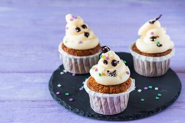 Carrot cupcakes with ghost cream decor and sprinkles on a slate board on a purple wooden background. Happy Halloween concept.