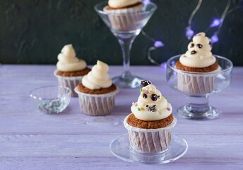 Carrot cupcakes with ghost cream decoration and sprinkles in a glass bowl on a purple wooden background. Halloween concept.