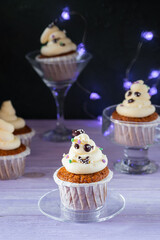 Carrot cupcakes with ghost cream decoration and sprinkles in a glass bowl on a purple wooden background. Halloween concept.