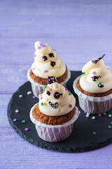 Carrot cupcakes with ghost cream decor and sprinkles on a slate board on a purple wooden background. Happy Halloween concept.