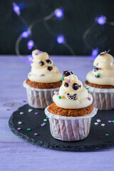 Carrot cupcakes with ghost cream decor and sprinkles on a slate board on a purple wooden background. Happy Halloween concept.