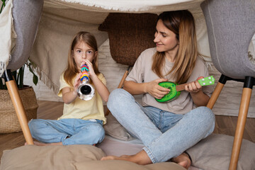 childhood and hygge concept - happy little girl with mother with musical instruments playing music in kids tent at home.
