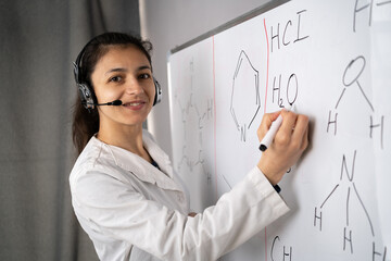 Portrait of female teacher with whiteboard background. Chemistry teacher with headset having online class and showing molecular model on flip chart, looking at camera