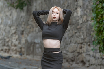 Slender blonde woman wears black tight-fitting clothes with her hands behind head and looks into camera on the street