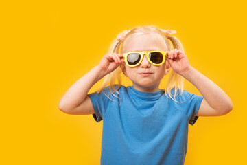 Studio portrait of preschool girl in sunglasses and blue T-shirt on yellow background. Little girl with blonde hair and two ponytails