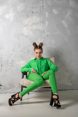 Slender girl with evening makeup and hairstyle poses on chair in studio. Lady in green pantsuit and heels on gray background