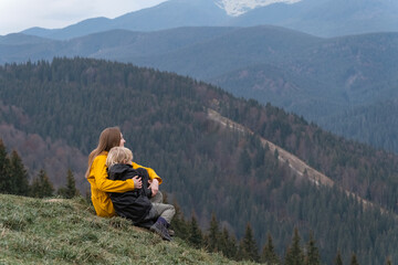 Fototapeta premium Mother and son rest and sit in an embrace on mountains background. Young mother with son sit on hillside