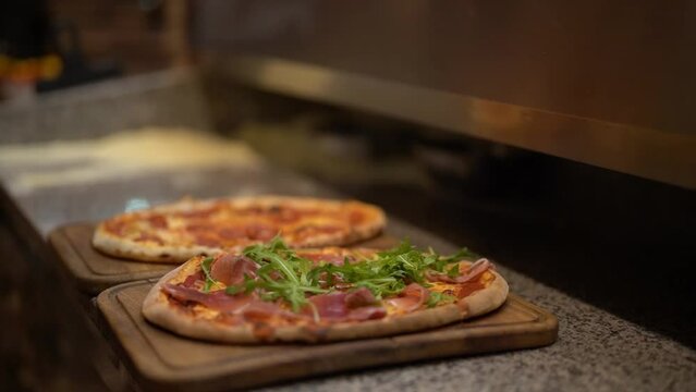 High angle of anonymous chef adding fresh green rucola to appetizing baked pizza with prosciutto served on wooden cutting board in kitchen of pizzeria