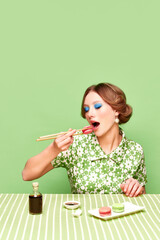 Pretty, stylish, young girl eating sweet macarons with chopsticks against green studio background. Soy sauce taste. Food pop art photography. Concept of retro style, creative vision, imagination.