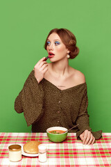 Pretty young girl with bright makeup eating vegetable soup against green studio background. Dinner time. Food pop art photography. Concept of retro style, creative vision, imagination.