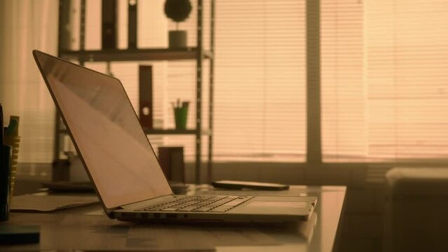 Empty workplace in a dark evening office. An open laptop, mobile phone and stationery lie on a table near a backlit window. The concept of office work, home online learning. Close up of silhouettes.
