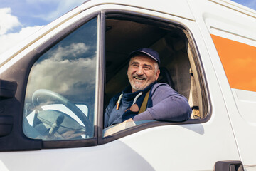 Portrait of a cheerful delivery driver looking out the window of the white cargo van vehicle,...