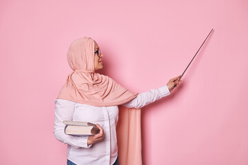 Inspired confident middle aged Arab Muslim woman, a school teacher or university professor explaining lesson, holding textbooks and pointing at copy space on pink background with a pointer. Education
