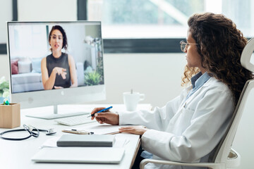 Beautiful young doctor woman doing videocall with patient in computer while taking some notes in the hospital consultation