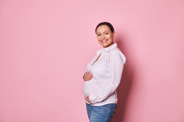 Isolated portrait pink background. Multi ethnic pregnant woman, touching her belly, enjoying first baby kicks, smiles looking at camera. Pretty gravid female expecting baby. Healthy pregnancy concept