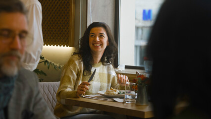 Young woman sits at the table in modern cafe, eats delicious food, works on freelance in laptop. Female freelancer has dinner in restaurant, talks to waiter. Concept of remote work and public catering