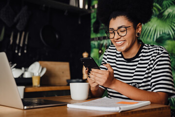 African woman drinking coffee and using smartphone while sitting with laptop in kitchen