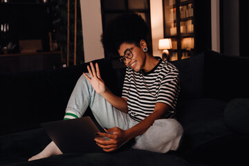 Cheerful african woman making video call via laptop and waving while sitting on sofa