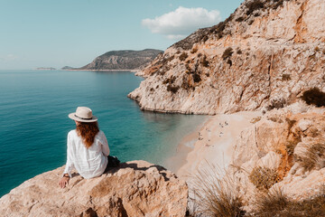 woman wearing a hat sitting on the rock watching beautiful sandy beach below.