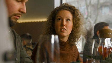 Middle aged woman talks with friends, watches menu and makes order. Waiter serves guest in cafe with modern design. Wine glasses and flowers stands on table. Concept of public eating. Portrait view.