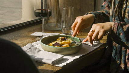Close up shot of female guest sitting near window, cutting food on pieces and tasting gourmet dish in restaurant with modern kitchen. Wine glass stands on table. Concept of public eating and catering.